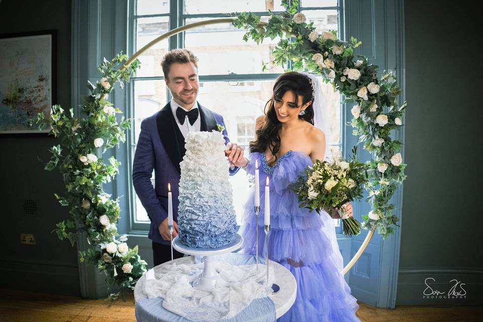 A bride and groom wearing blue wedding outfits cutting into a blue wedding cake in front of a floral arch
