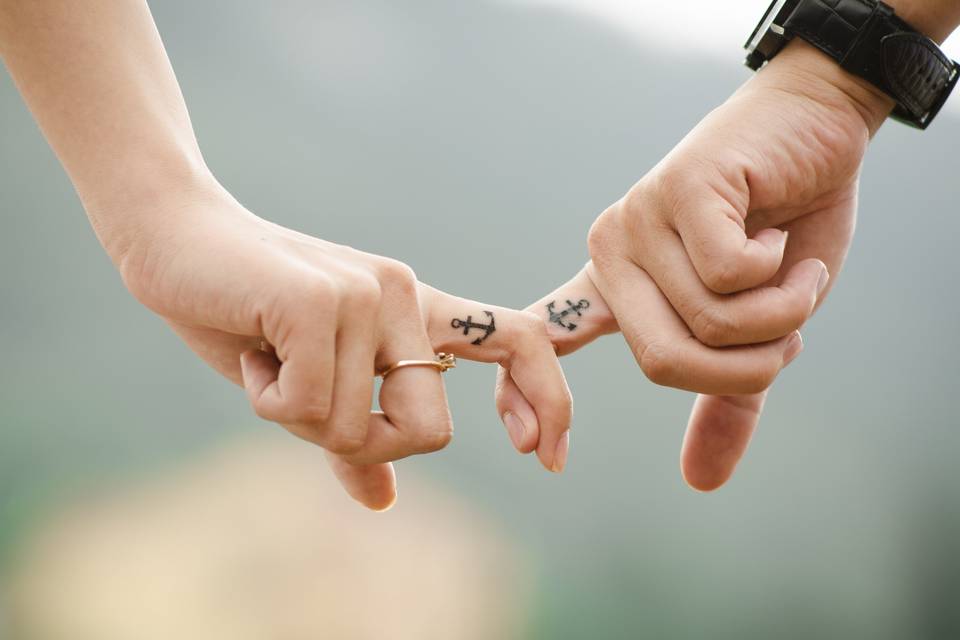 White woman with a gold diamond ring and anchor tattoo on her inner finger linking fingers with a white man wearing a black watch with a matching anchor tattoo on his inner finger