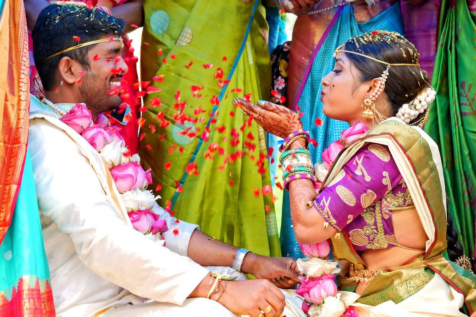 Hindu bride and groom sitting opposite one another wearing flower garlands with the bride blowing petals in the groom's face and the newlyweds surrounded by family in colourful outfits