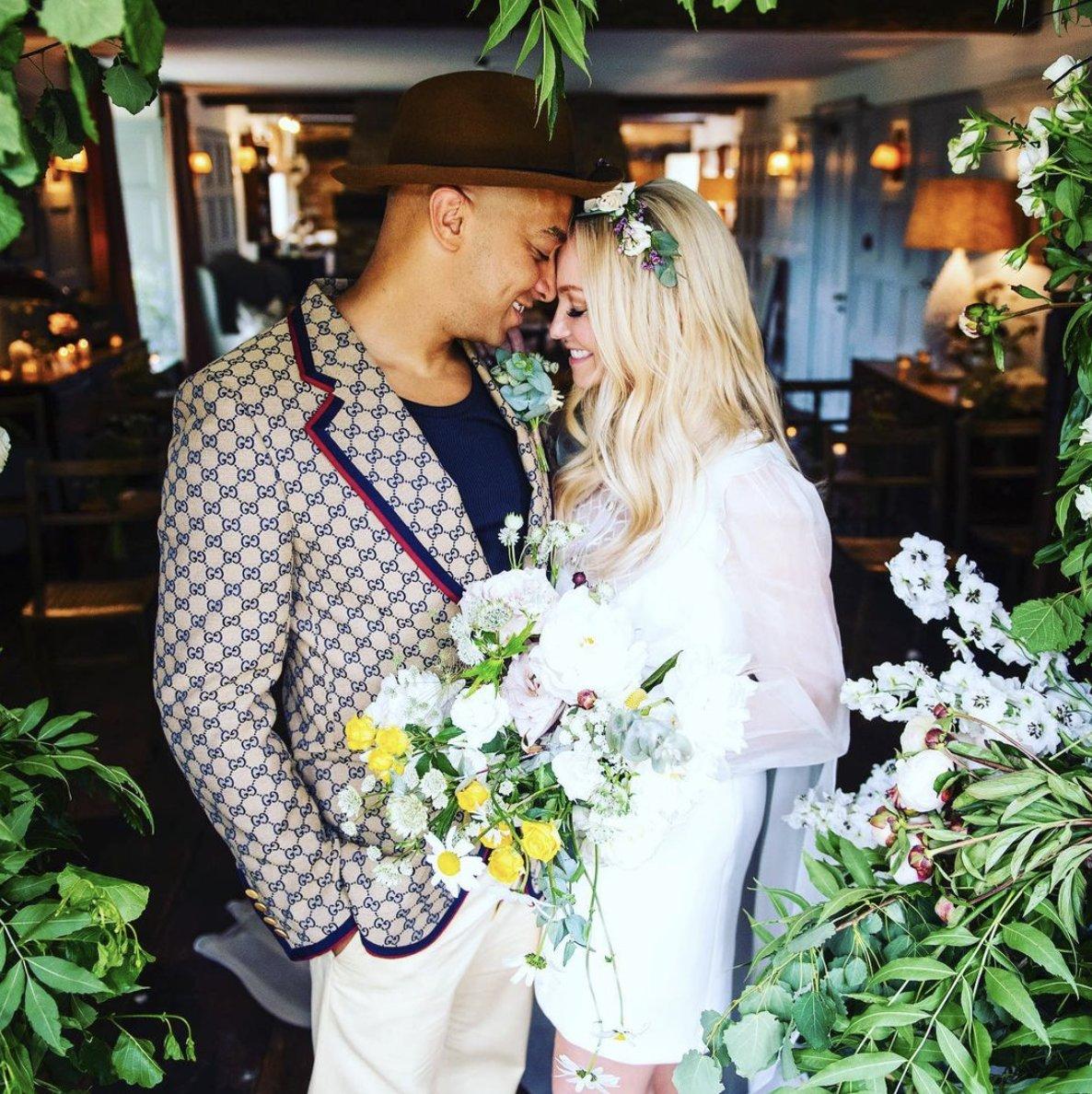 Emma Button and Jade Jones touching foreheads under a flower arch with Emma wearing a flower crown and short white dress with a yellow and bouquet and Jade in a brown trilby hat, patterned brow and beige jacket and beige chinos