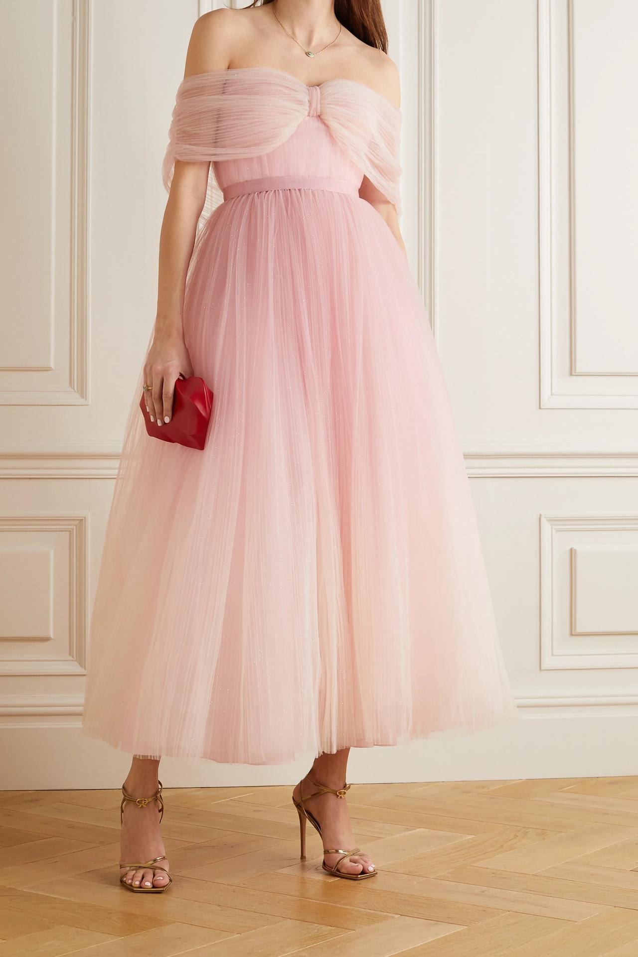 White woman wearing an off the shoulder light pink glittery tulle gown with a small red clutch bag