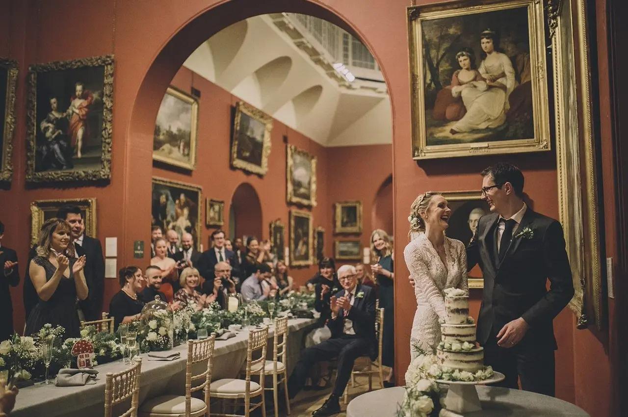 Laughing newlyweds cut the wedding cake in a gallery as their guests applaud