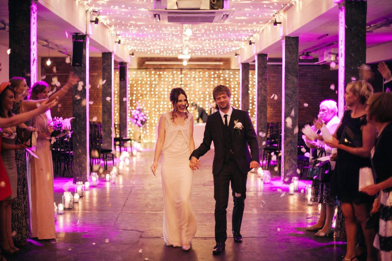 Bride and groom walk down the candle lit aisle with fairy lights and purple lighting
