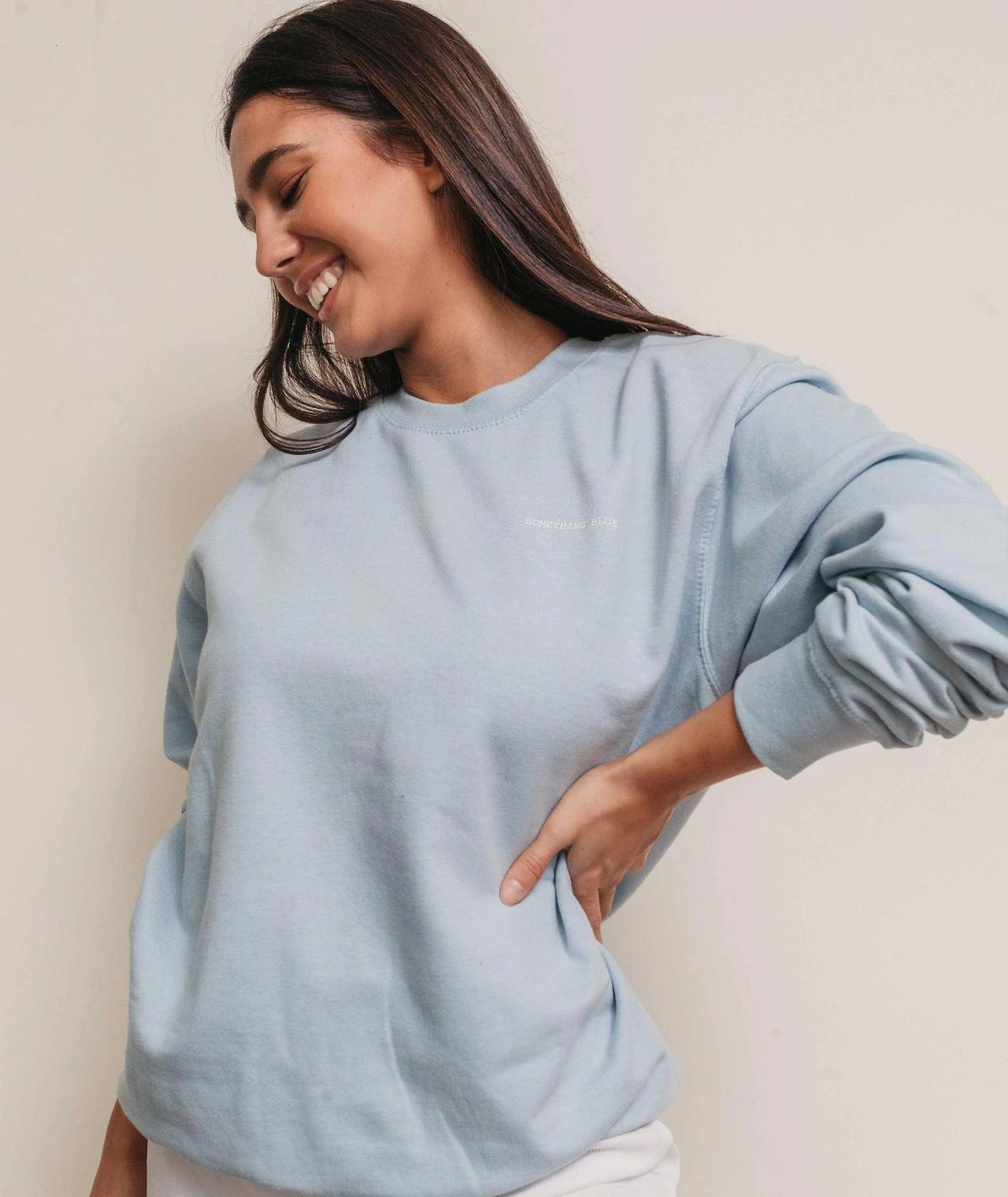 Tanned woman with brown hair laughing wearing a light blue jumper with something blue embroidered onto the front