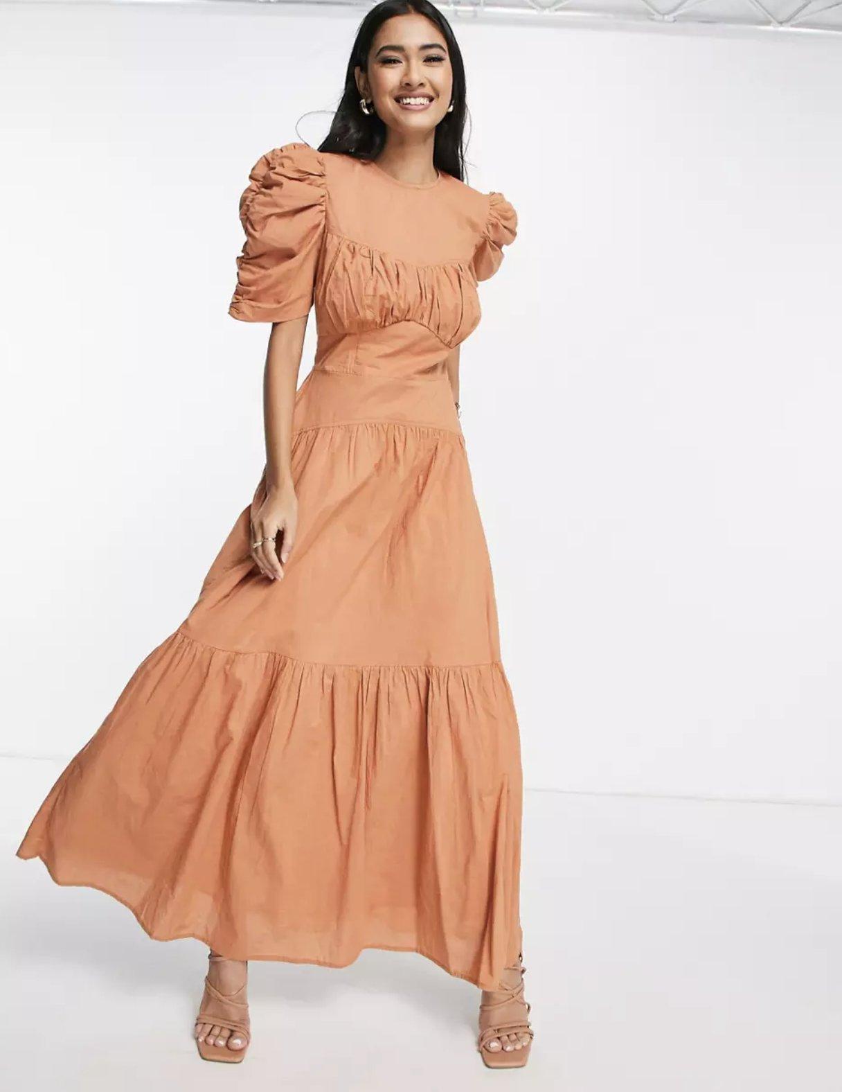 Image of a model mid-stride wearing a rust-colored dress with ruched sleeves and a tiered flowing skirt