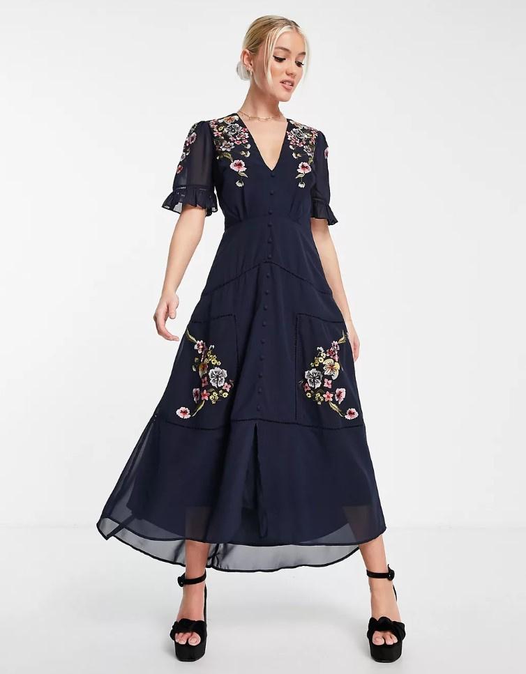 Image of a model wearing a navy midi dress with ruffled sleeves, floral embroidery, button front details