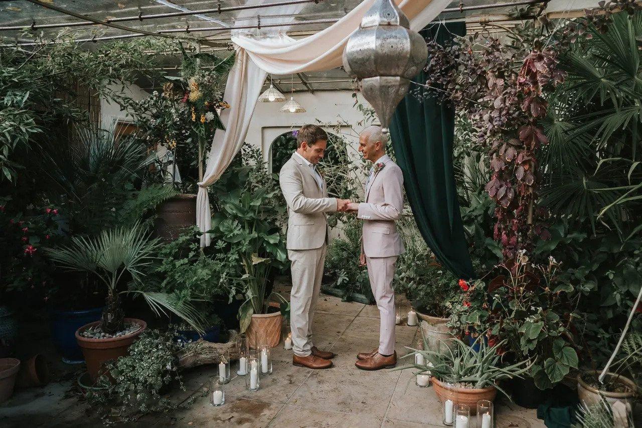 An LGBTQ+ couple stand at a greenery surrounded wedding altar holding hands as they exchange vows