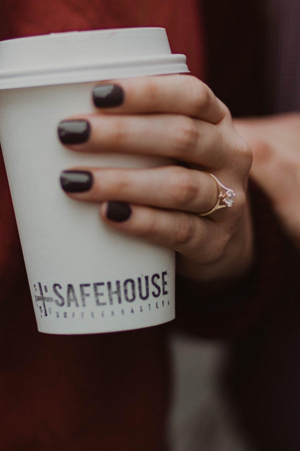 Close up of white woman's hand with a black manicure holding a white coffee cup wearing a gold ring with a pyramid diamond at its centre