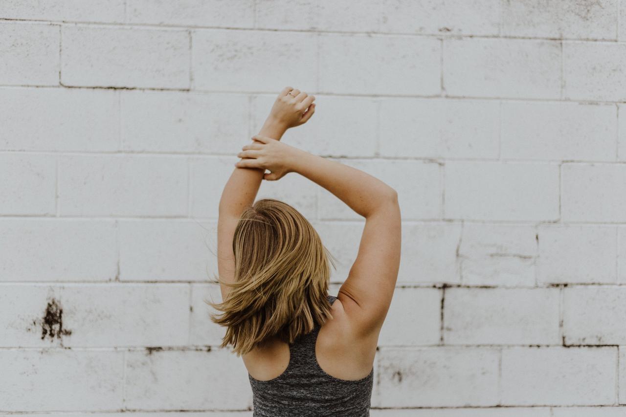 White woman with blonde hair dancing in a grey top facing a white brick wall