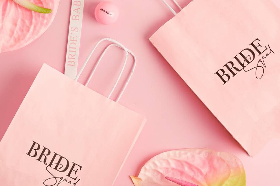 Paper bags with 'Bride Squad' written on them. They are lying on a pink background with a hair clip and a nail file.