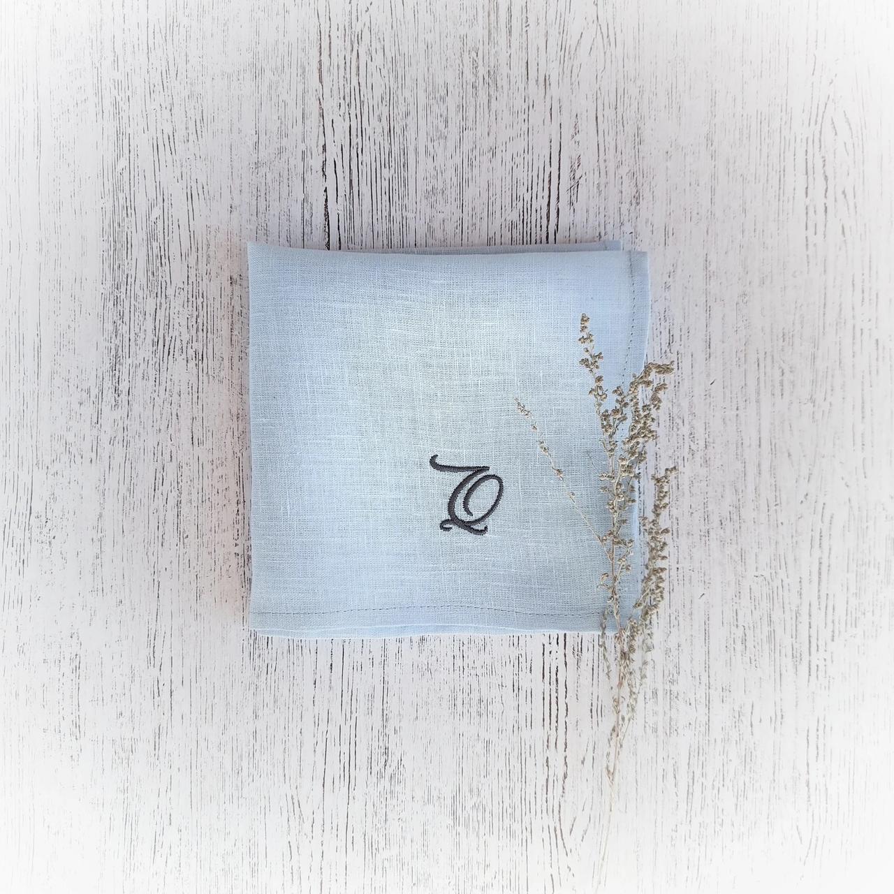 Light blue linen handkerchief with a 'Q' embroidered in navy thread next to a spring of dried flowers on a light wooden tablecloth