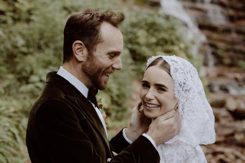 Lily Collins wearing a hooded lace long sleeved wedding dress smiling at the camera opposite her new husband Charlie McDowell in a dark velvet suit cupping Lily's face