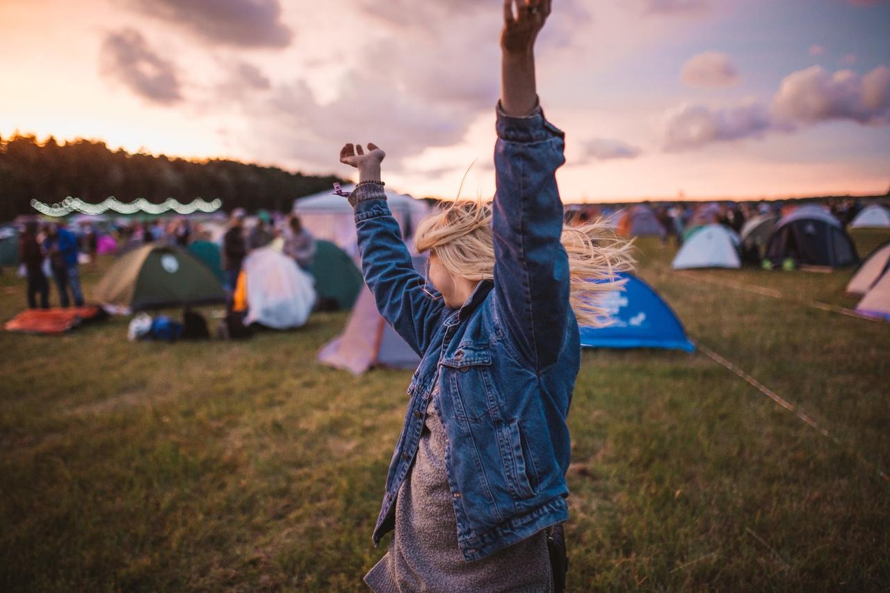 Blonde woman in a denim jacket dancing in the sun in a field full of coloured tents