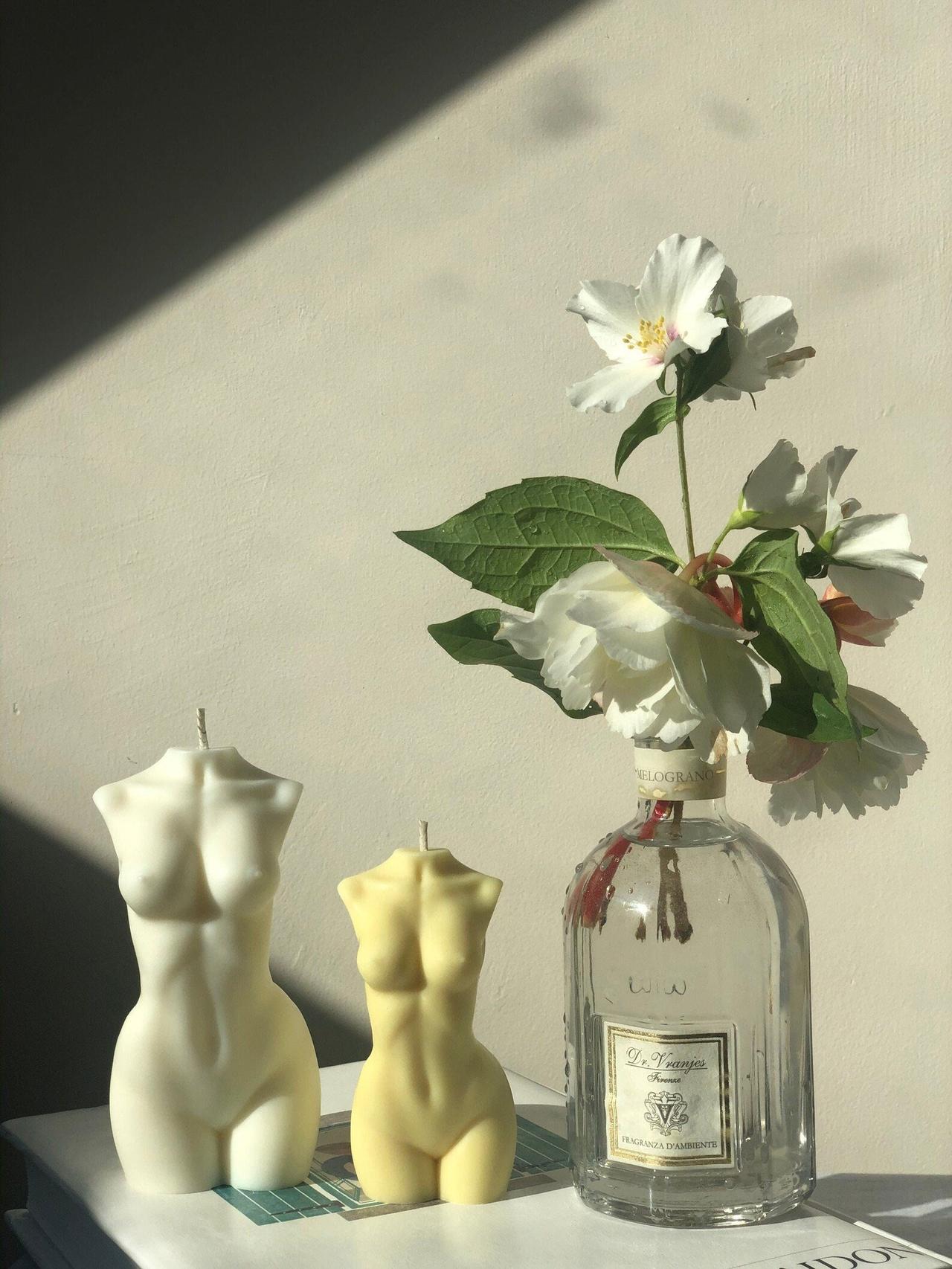 Two candles in the shape of a Venus bust on a table alongside a vase of flowers. One is large, one is smaller