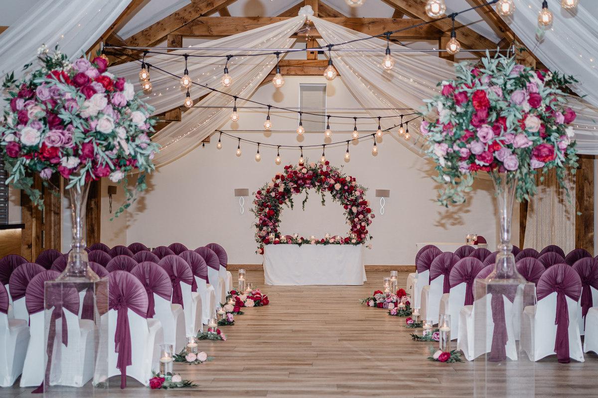 Interior of a barn wedding venue set up for a ceremony with chair covers and sashes and a floral arch at the altar.