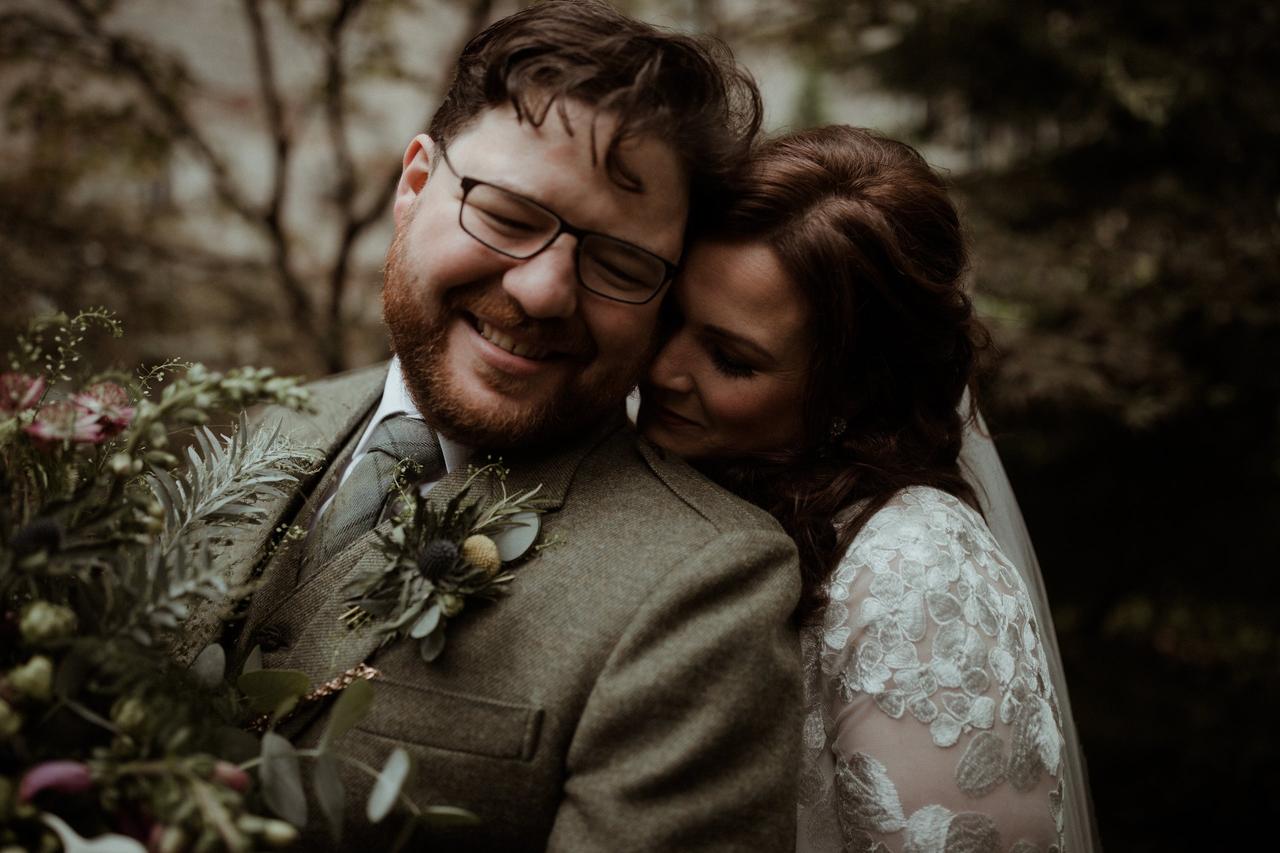 Laughing white groom with a beard and glasses wearing a light green tweed suit with a thistle buttonhole holding a foliage bouquet with his bride leaning on his shoulder wearing an embroidered floral white wedding dress