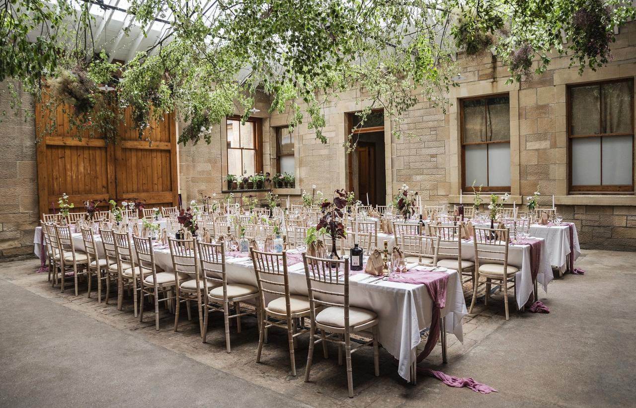 A coach house reception set up with greenery on the ceilings, natural light and long wedding tables.