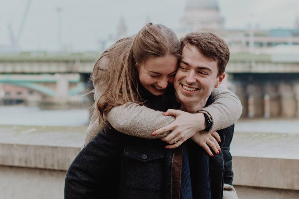 A smiling man carrying his new wife-to-be on his back in front of the London city skyline