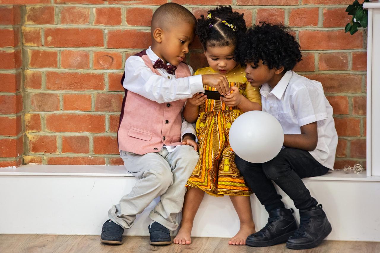 Two young boys and a little girl huddled in a corner looking at something the girl is holding. They are dressed in smart party clothes and one of the boys is holding a balloon.