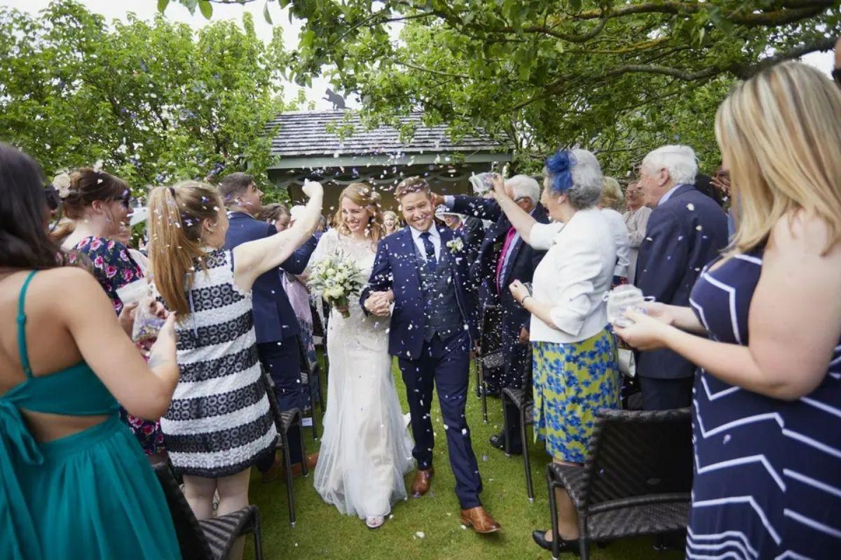 a bride and groom leaving their wedding ceremony as guests throw confetti over them at a summer garden wedding