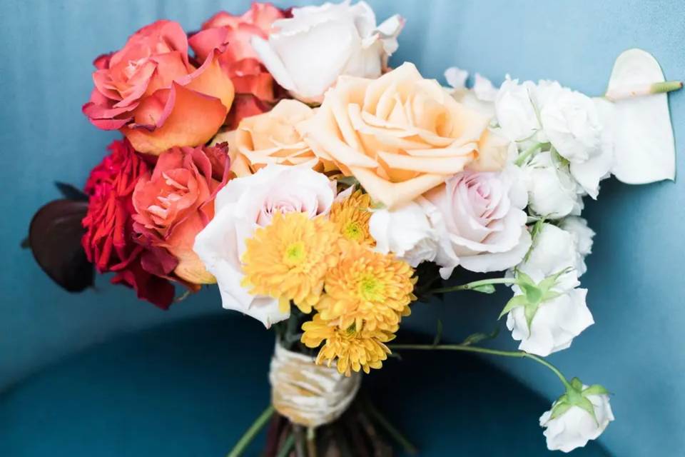 A bright coloured ombre wedding bouquet made up of a range of May wedding flowers sits on a light blue leather chair. The flower colours go from white to orange to red