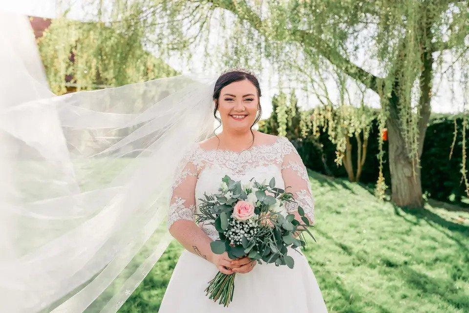 Bride holding a bouquet smiles at the camera outdoors while her veil blows in the wind
