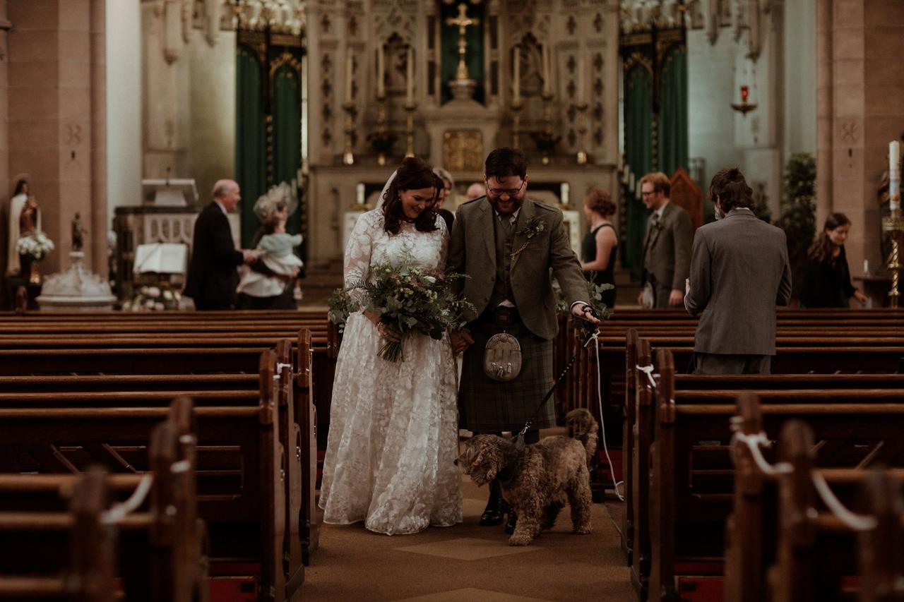 White bride and groom walking back down the aisle of a church with their dog on a lead in front of them and wedding guests in pews behind them