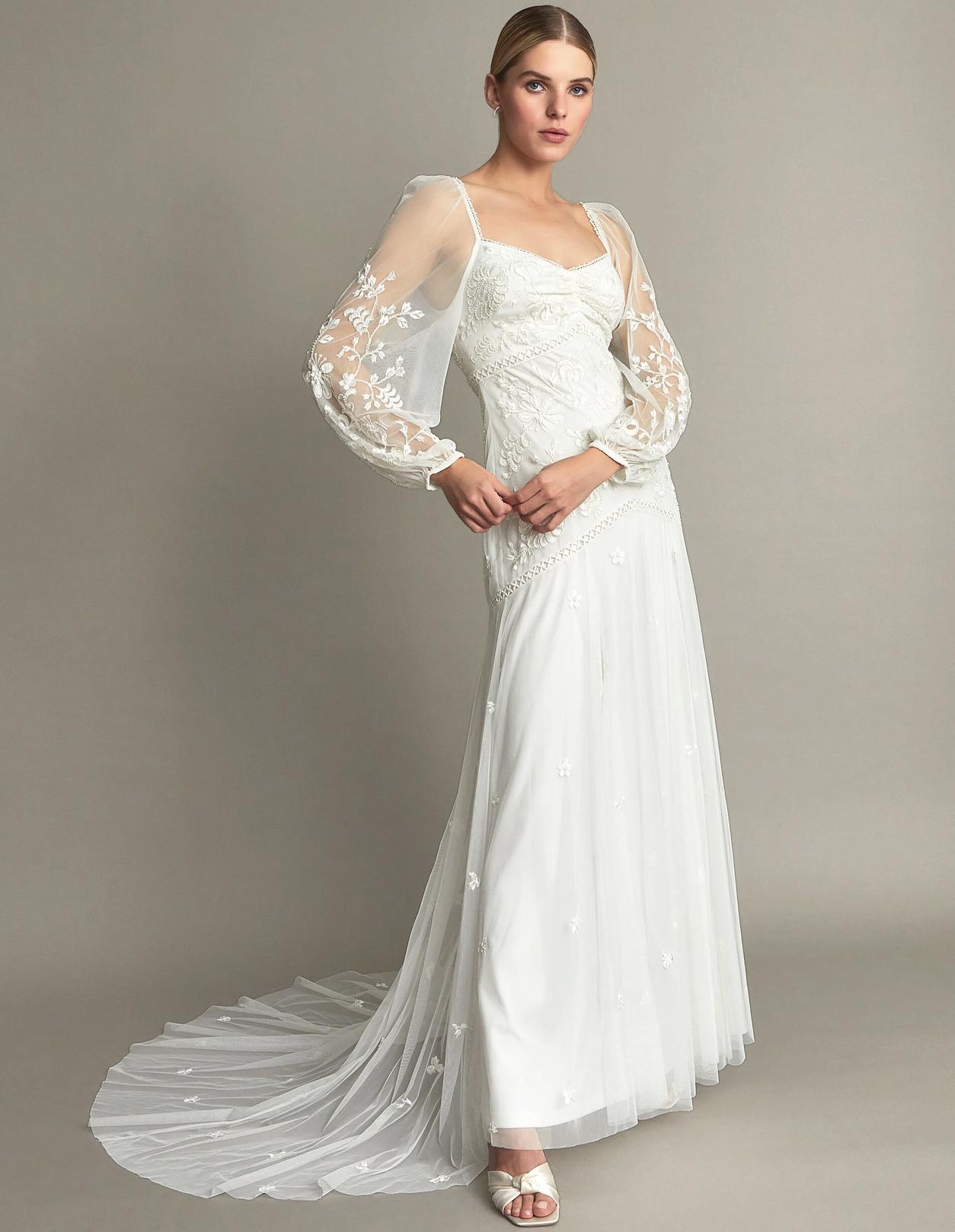 woman stands with one foot in front of the other, wearing a long sleeved floor length embroidered vintage style wedding dress