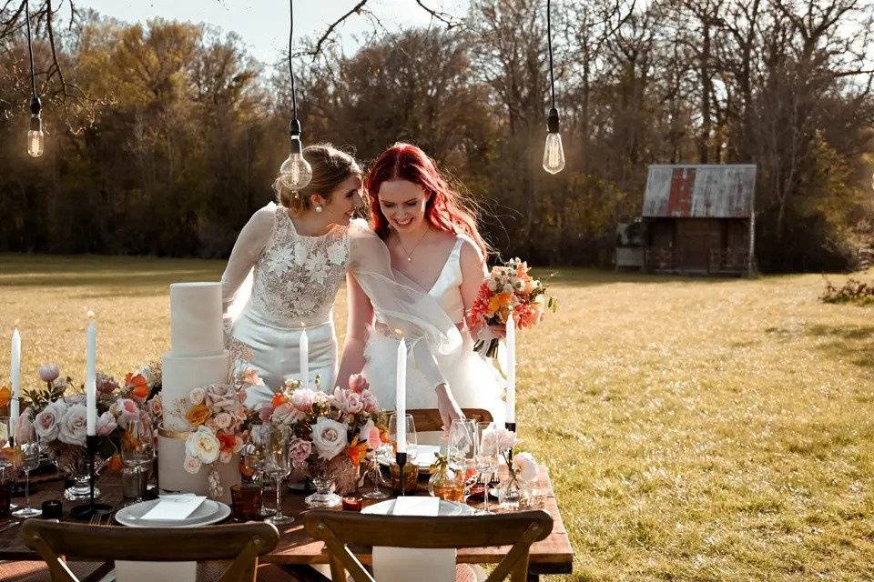 A candid picture of twi brides in front of an outdoor table laid with flowers and dinner candles with exposed bulbs hanging above