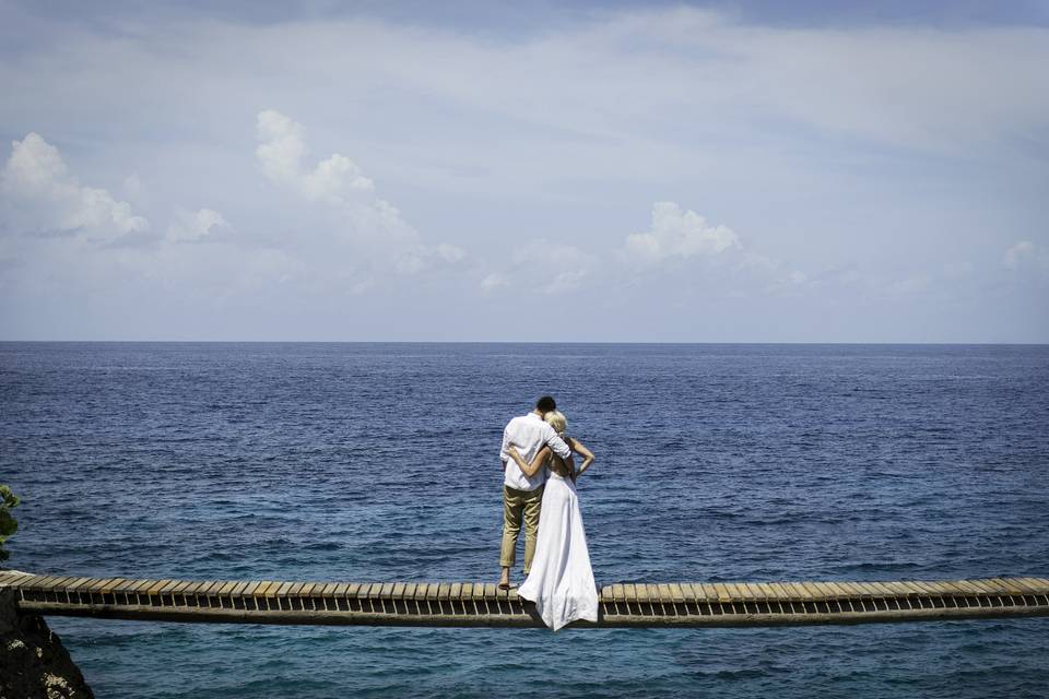 Embracing couple look at the ocean from a swinging bridge over the water.