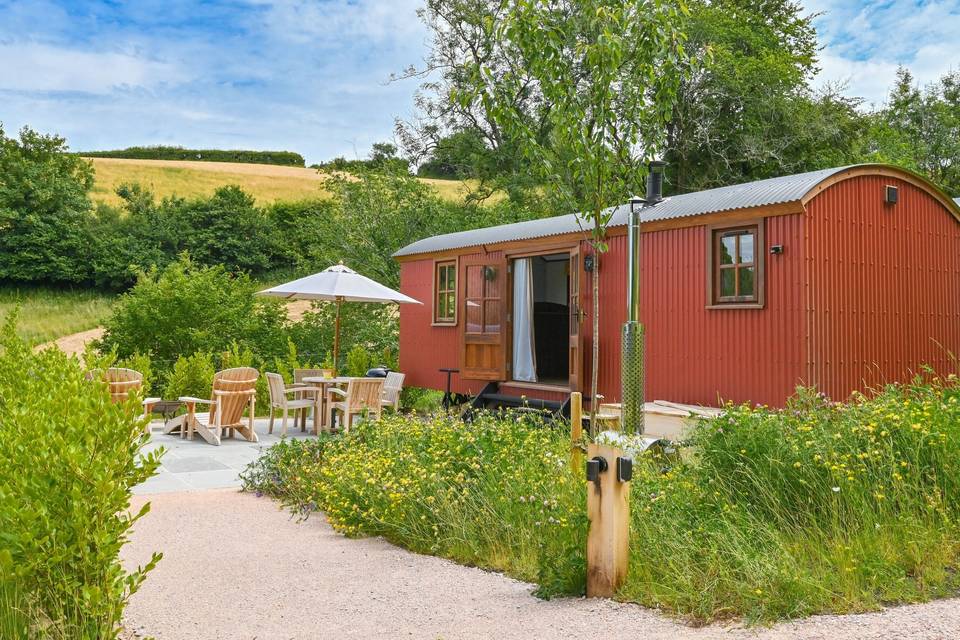 Outdoor view of a shepherd's hut mini moon accommodation in a flower strewn meadow with a seating area outdoors