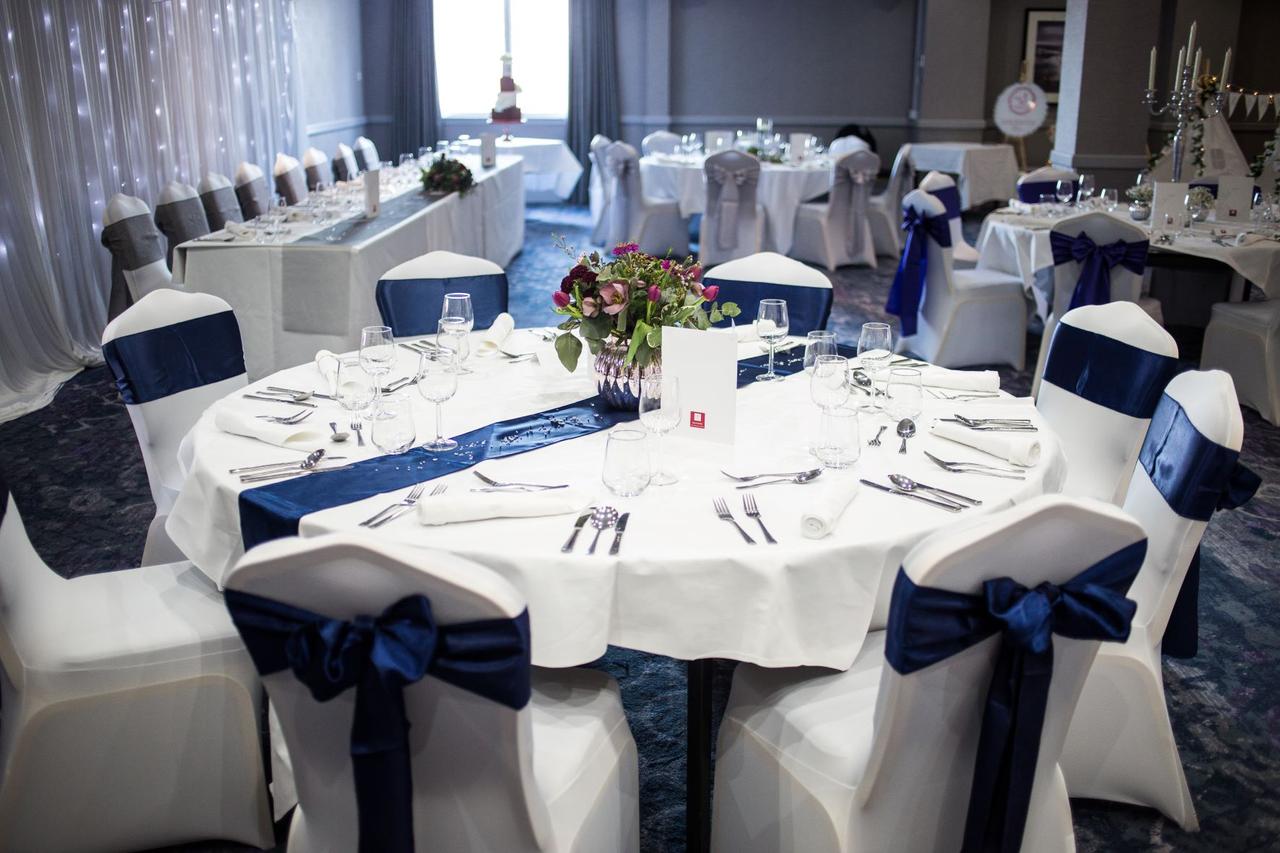 A hotel function room set up for a wedding breakfast with round tables and white chair covers with navy blue ribbon.