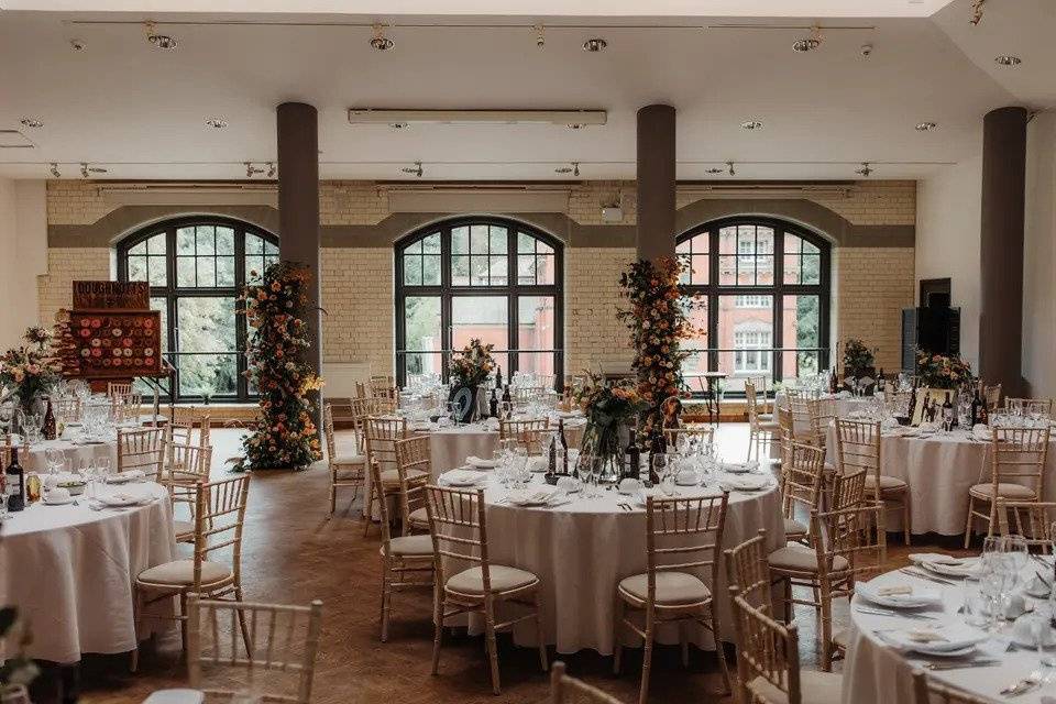 Room set for wedding meal with low ceiling, tiled walls, large windows, round white tables, wooden chairs and floral centrepieces