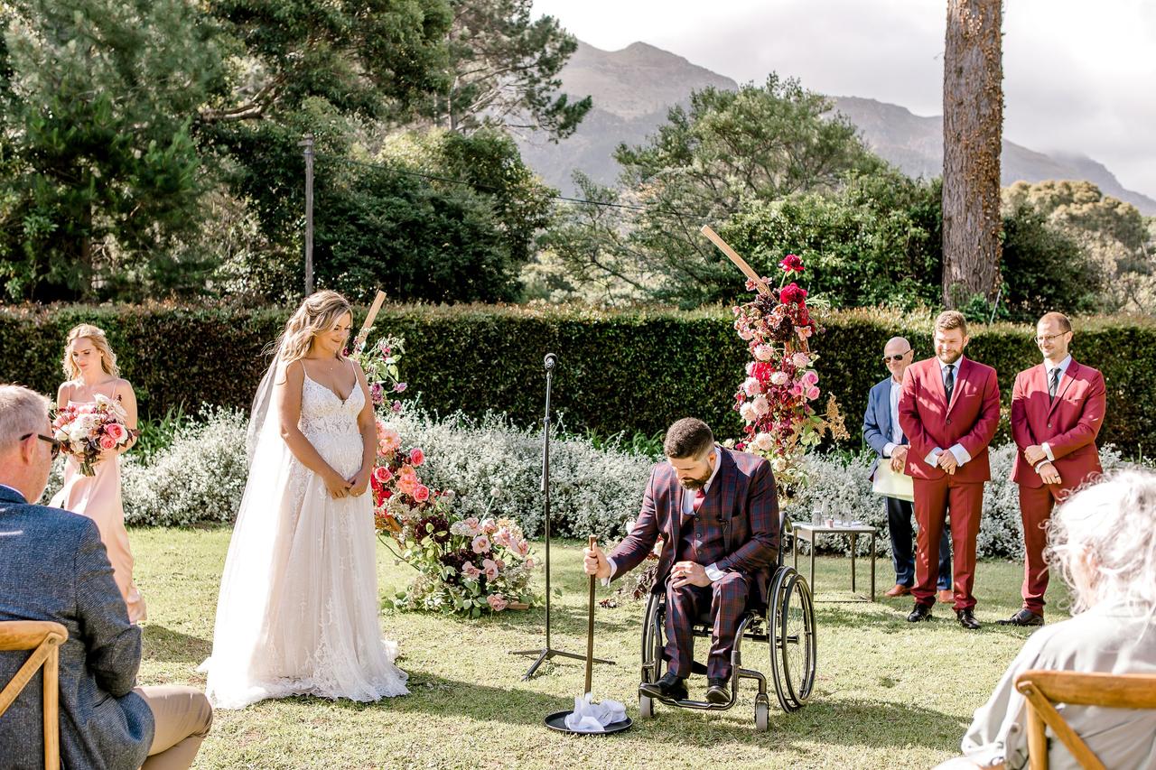 A groom sits and crushes a glass with a stick while his bride and their wedding guests watch on