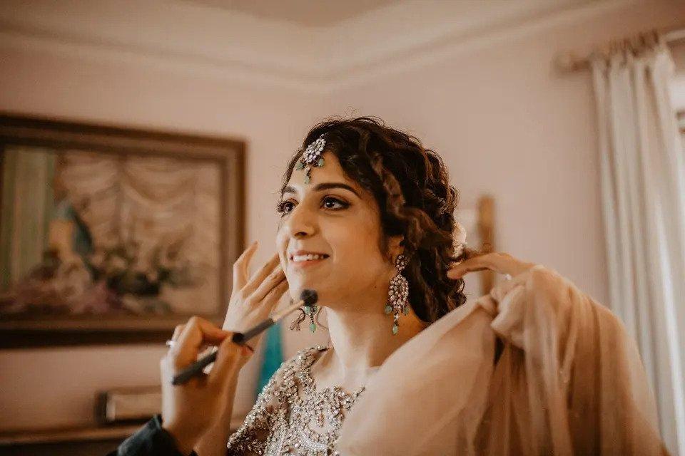 A bride dressed in traditional Indian wedding garments and accessories looks into the distance as an off-screen MUA adjusts her makeup