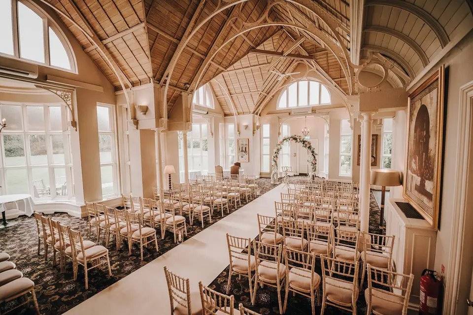 Room set up for wedding ceremony with vaulted wooden ceilings, floor-to-ceiling windows, a white carpet down the aisle, wooden chairs and a floral arch