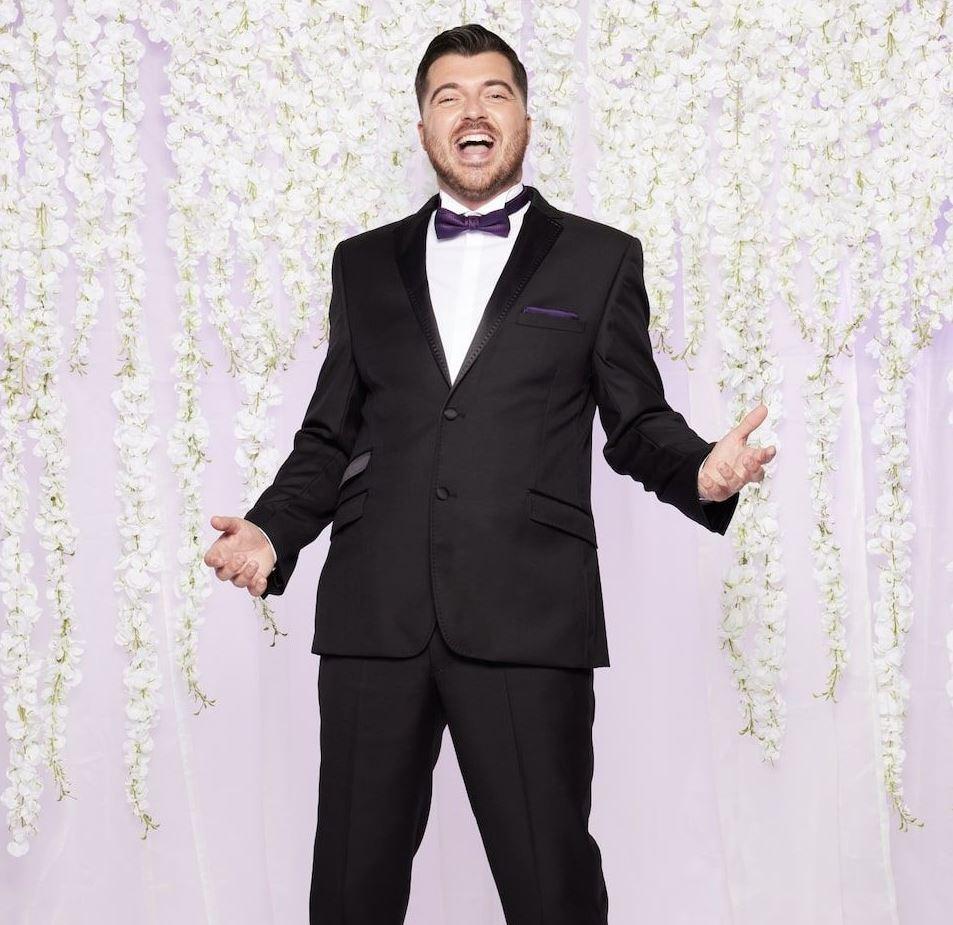 mafs groom luke laughs in a black tux with purple accents as he stands in front of a lilac backdrop with white hanging flowers