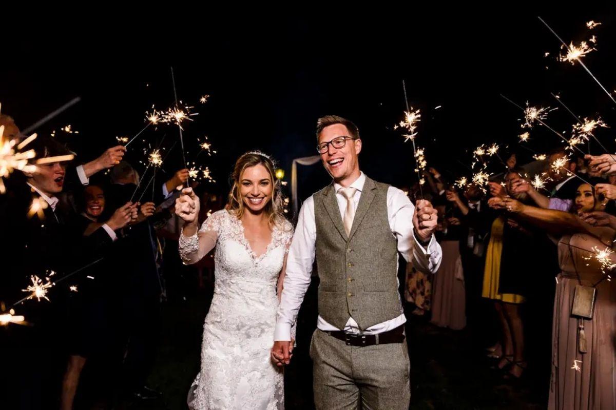 a bride and groom hand in hand waving sparklers with their guests doing the same at night time