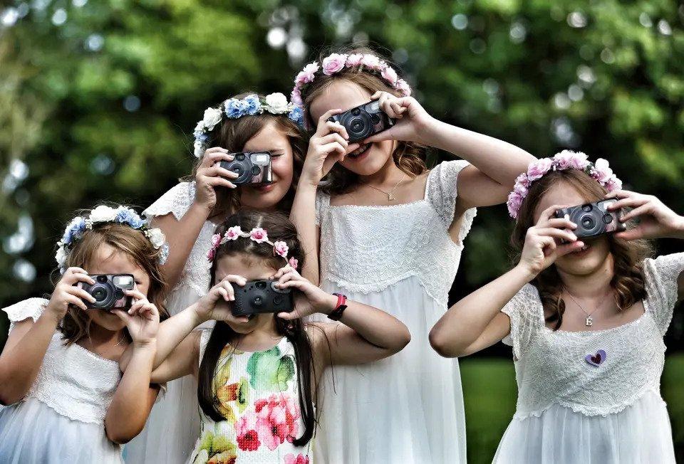 Group of flower girls in matching dresses and crowns taking pictures of the camera with disposable cameras