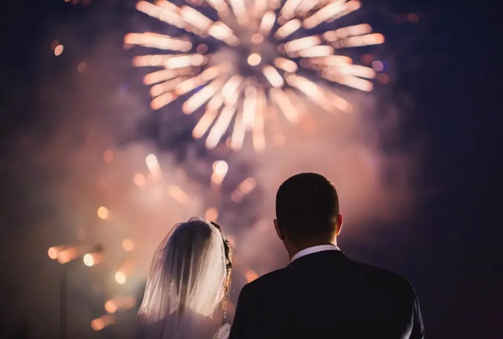 a shot over the shoulder of a bride and groom as they watch fireworks at their wedding