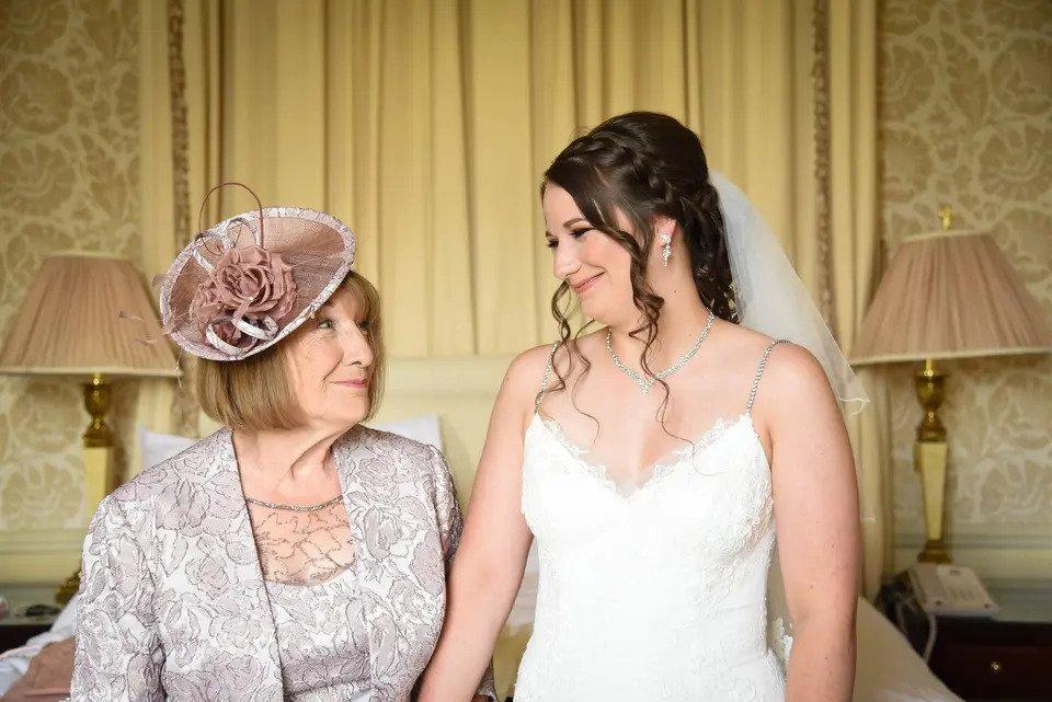 Bride and mother of the bride stand hand-in-hand and smile at each other