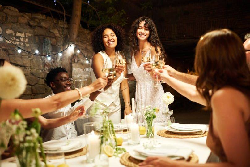 LGBTQ+ couple celebrating their wedding day with friends. The two women are dressed in white, smiling and raising a champagne toast.