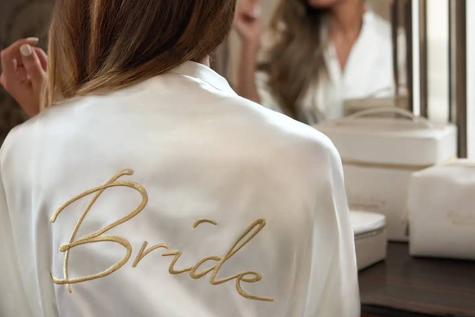 a woman wearing a bride dressing gown made from white satin with the word 'bride' embroidered on the back in gold
