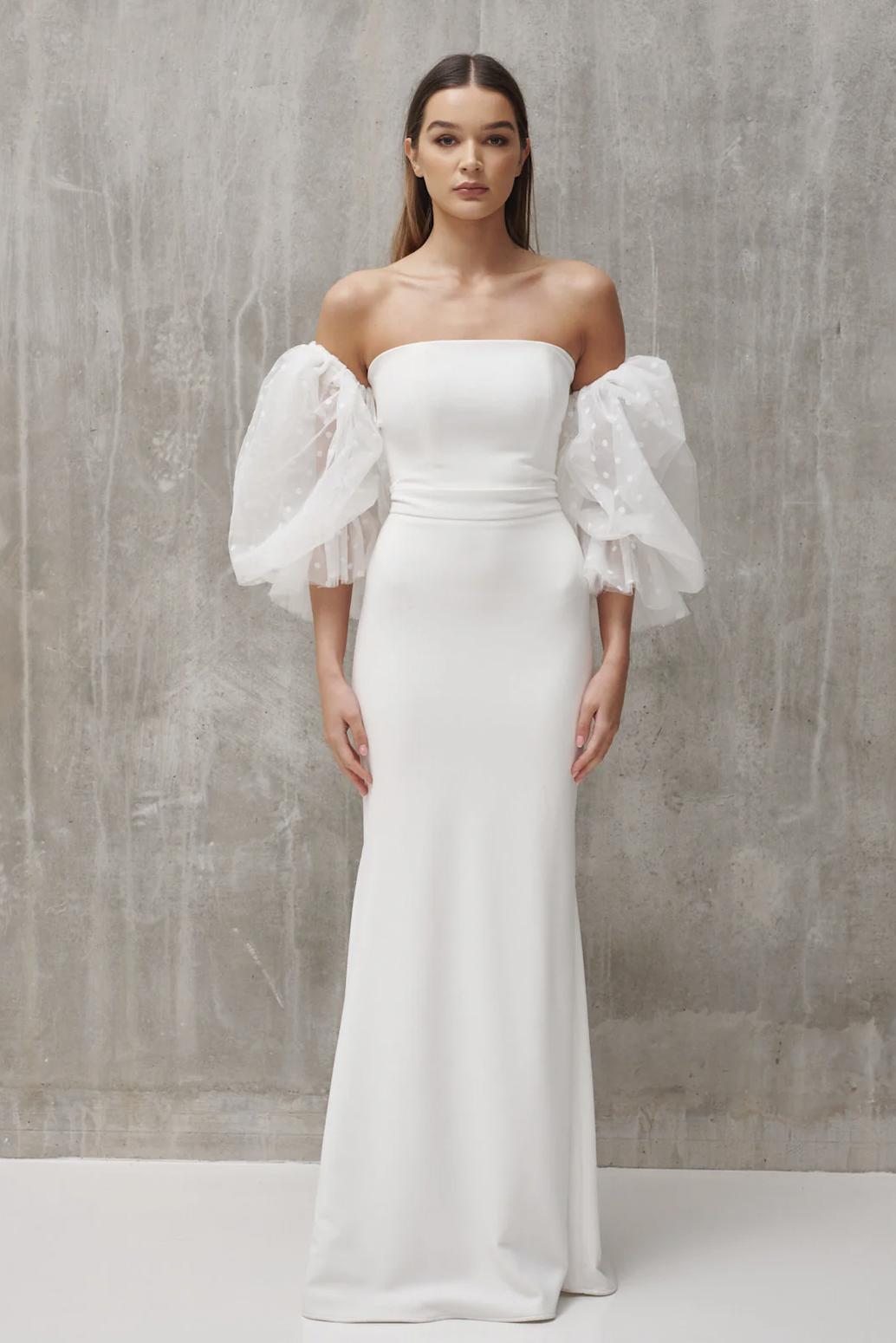 bride standing in front of grey wall wearing a form-fitting column dress in white with detachable puff sleeves in a polka dot fabric