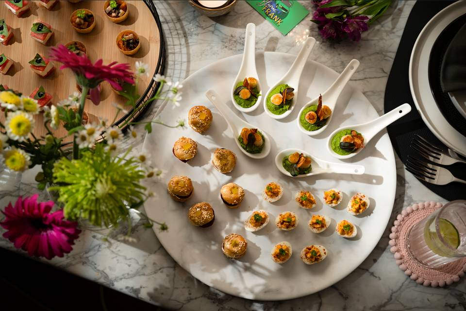 Two large plates of canapes displayed on a table dressed for a wedding, decorated with spoons holding elegant canapes, egg sliders and mini hamburgers