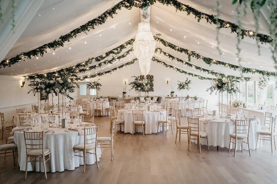 Room set for wedding reception with marquee style roof, fairy lights, green foliage, hanging chandelier, round white tables, Chiavari chairs and floral centrepieces