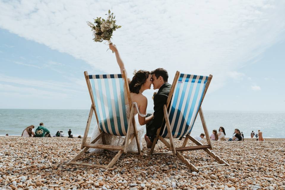 A bride and groom in their wedding attire sitting in blue striped deckchairs bringing their faces close together ready to kiss on a pebble beach