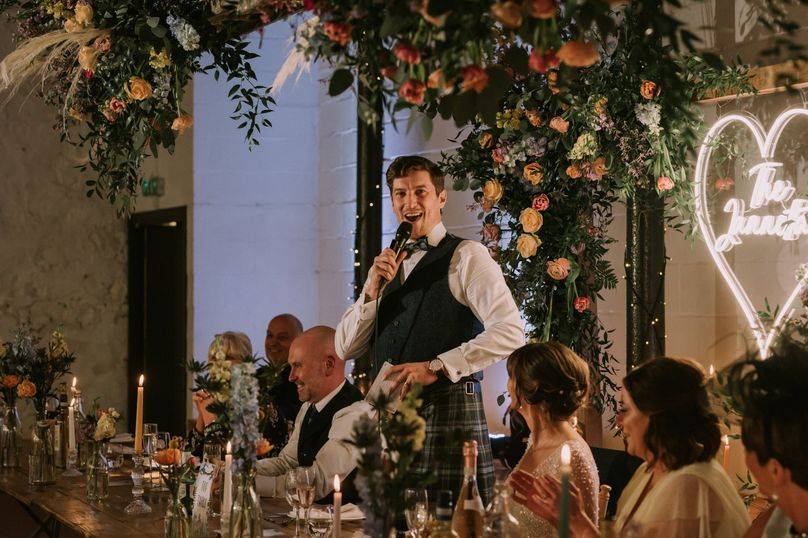 Best Man telling a funny joke during his wedding speech at a traditional wedding. All the guests are laughing and smiling at the candlelit tables.