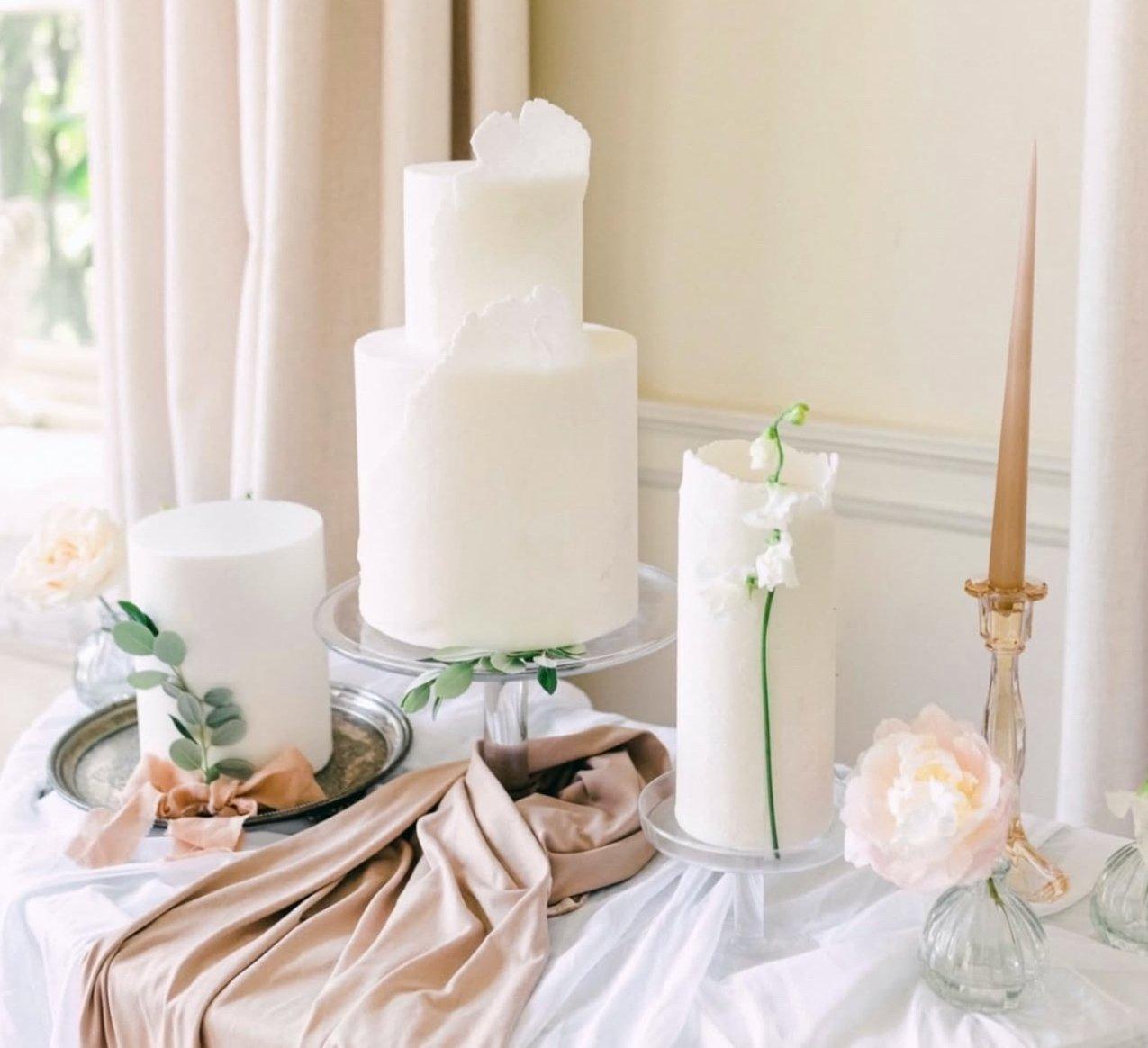 A trio of white iced wedding cakes arranged on a table with silks, candles and eucalyptus.