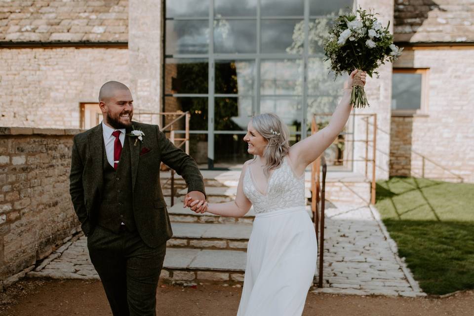 A jubilant bride and groom leaving their wedding venue grinning at each other, hand in hand, whilst the bride holds her bouquet aloft in the air in triumph